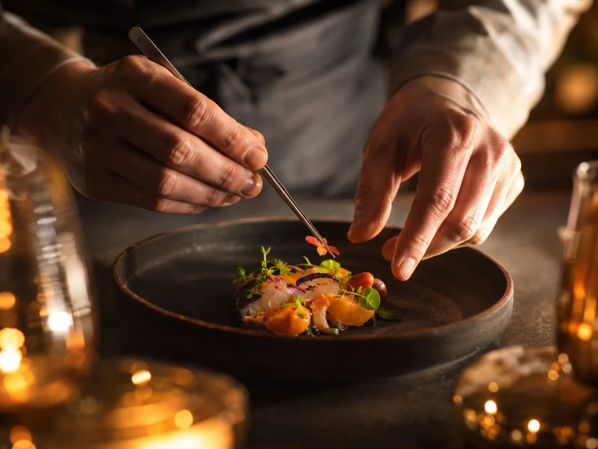 A chef&rsquo;s hands plating a course with tweezers by candlelight