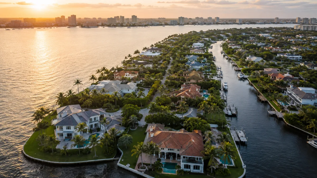 Waterfront residences and their gardens seen from the air at golden hour, canals threading between them toward a distant skyline